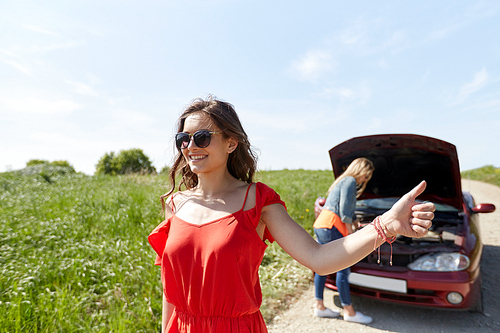 road trip, transport, travel, gesture and people concept - young women with broken car showing hitchhiking gesture asking for help at countryside