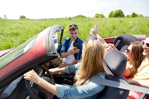 leisure, road trip, travel and people concept - happy friends driving in cabriolet car and taking picture by smartphone
