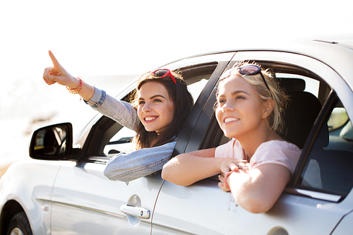 summer vacation, holidays, travel, road trip and people concept - happy teenage girls or young women sitting in car at seaside and pointing finger to something