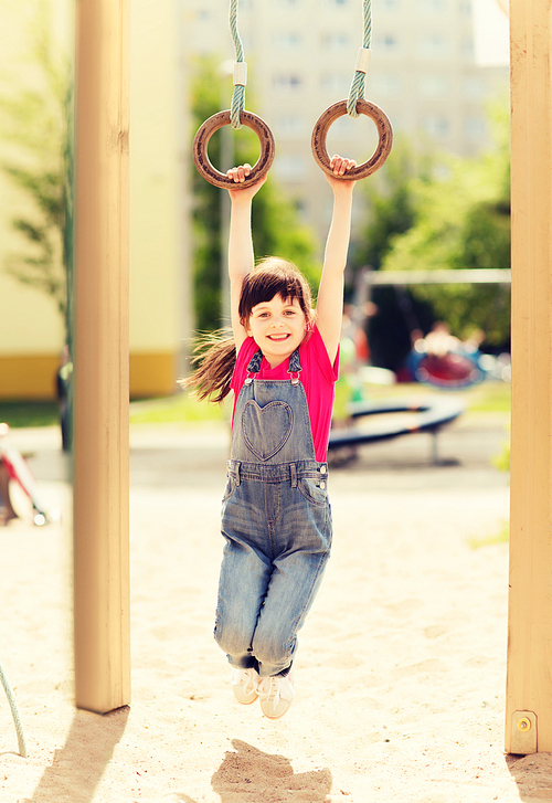 summer, childhood, leisure and people concept - happy little girl hanging on gymnastic rings at children playground