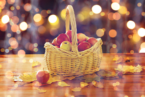 harvest, season, autumn and fruits concept - close up of wicker basket with ripe red apples and leaves on wooden table over holidays lights
