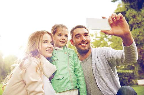 family, childhood, technology and people concept - happy father, mother and little daughter taking selfie by smartphone in park