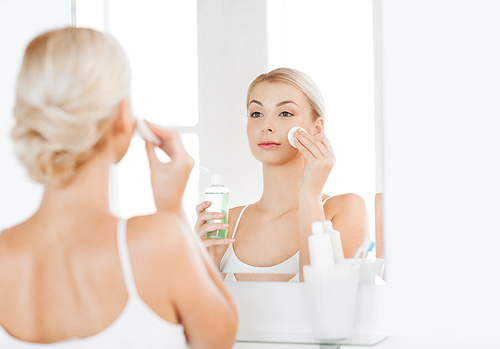 beauty, skin care and people concept - smiling young woman applying lotion to cotton disc for washing her face at bathroom