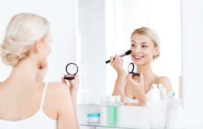 beauty, make up, cosmetics, morning and people concept - smiling young woman applying blush with makeup brush and looking to mirror at home bathroom