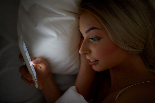 technology, internet, communication and people concept - happy smiling young woman texting on smartphone in bed at home bedroom at night