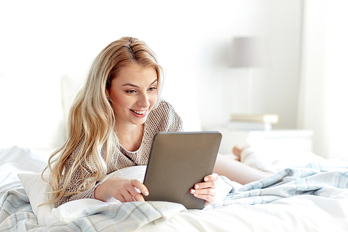 technology, internet and people concept - happy young woman lying in bed with tablet pc computer at home bedroom