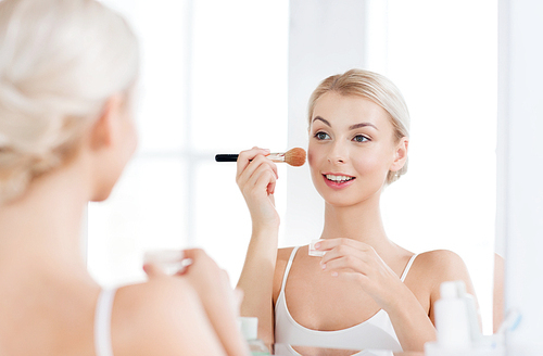 beauty, make up, cosmetics, morning and people concept - smiling young woman makeup brush and powder foundation looking to mirror at home bathroom