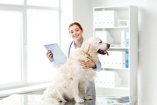 medicine, pet, animals, health care and people concept - happy veterinarian or doctor with golden retriever dog and clipboard at vet clinic