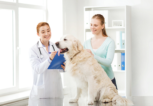 medicine, pet, animals, health care and people concept - happy woman with golden retriever dog and veterinarian doctor writing to clipboard at vet clinic