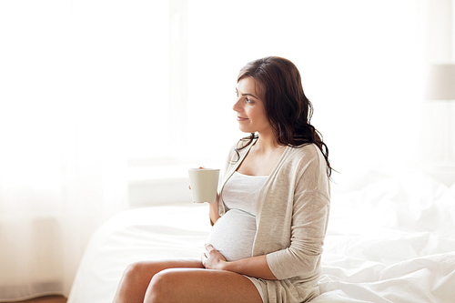 pregnancy, drinks, rest, people and expectation concept - happy pregnant woman with cup drinking tea sitting on bed at home bedroom