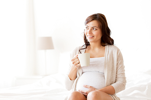 pregnancy, drinks, rest, people and expectation concept - happy pregnant woman with cup drinking tea sitting on bed at home bedroom