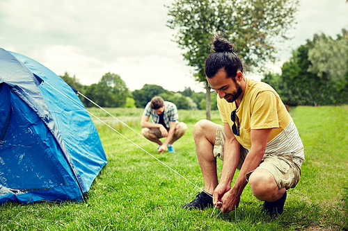 camping, travel, tourism, hike and people concept - smiling friends setting up tent outdoors