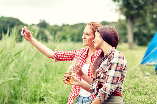 camping, travel, tourism, hike and people concept - happy young women with glass bottles drinking cider or beer and taking selfie by smartphone at camping