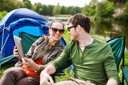 travel, hiking, technology, tourism and people concept - smiling couple with tablet pc computer sitting on chairs at camping tent