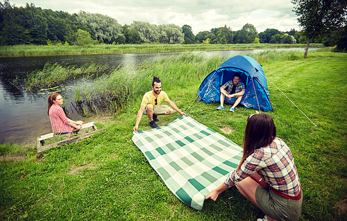 camping, travel, tourism, hike and people concept - happy friends with tent laying picnic blanket at campsite on river bank