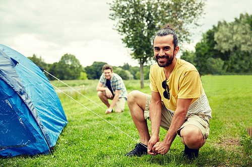 camping, travel, tourism, hike and people concept - smiling male friends setting up tent outdoors