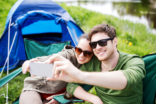 travel, hiking, technology, tourism and people concept - smiling couple of travelers taking selfie by smartphone at camping