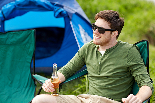 camping, travel, tourism, hike and people concept - happy young man drinking beer at campsite tent