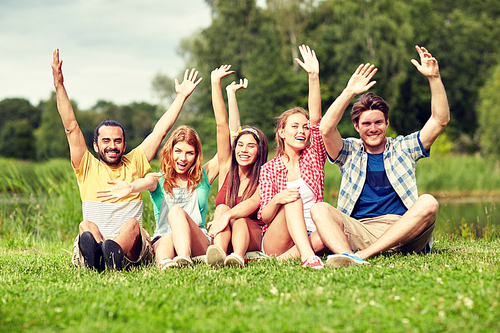 friendship, leisure, summer and people concept - group of smiling friends sitting on grass and waving hands outdoors