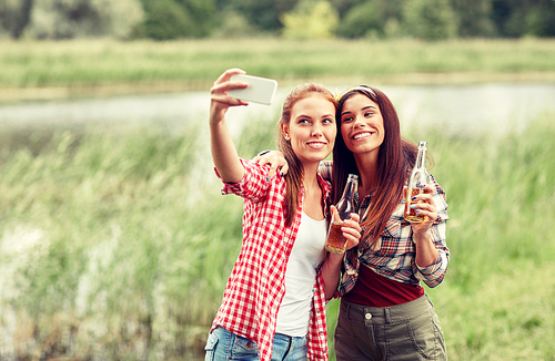 camping, travel, tourism, hike and people concept - happy young women with glass bottles drinking cider or beer and taking selfie by smartphone outdoors