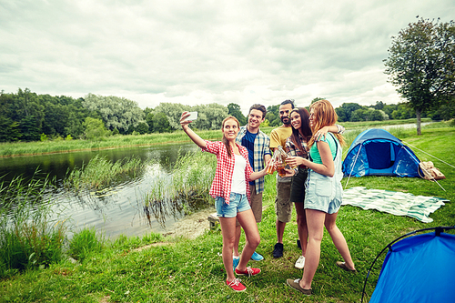 camping, travel, tourism, hike and people concept - happy friends with glass bottles drinking cider or beer and taking selfie by smartphone at camping