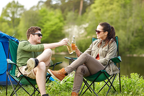 camping, travel, tourism, hike and people concept - happy couple clinking beer bottles at campsite tent