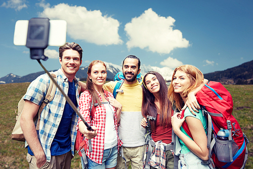 technology, travel, tourism, hike and people concept - group of smiling friends walking with backpacks taking picture by smartphone on selfie stick over natural background