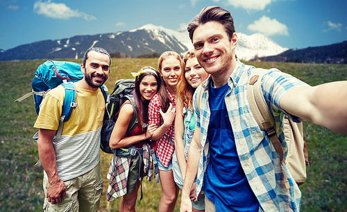 technology, travel, tourism, hike and people concept - group of smiling friends walking with backpacks taking selfie by smartphone or camera in over natural background