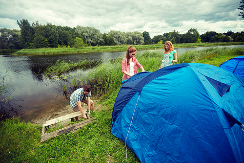 camping, travel, tourism, hike and people concept - group of smiling friends setting up tent outdoors