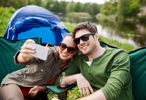 travel, hiking, technology, tourism and people concept - smiling couple of travelers taking selfie by smartphone at camping