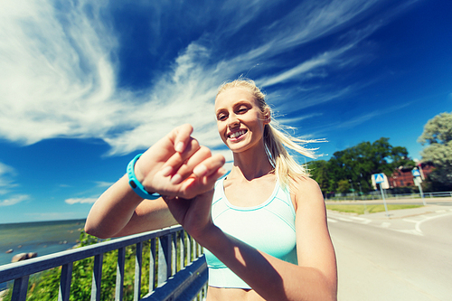 fitness, sport, people, technology and healthy lifestyle concept - smiling young woman with heart rate watch and earphones exercising outdoors