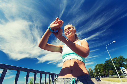 fitness, sport, people, technology and healthy lifestyle concept - smiling young woman with heart rate watch and earphones exercising outdoors