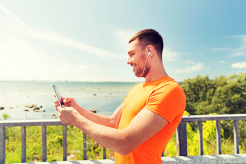 fitness, sport, people, technology and healthy lifestyle concept - smiling young man with smartphone and earphones listening to music at summer seaside