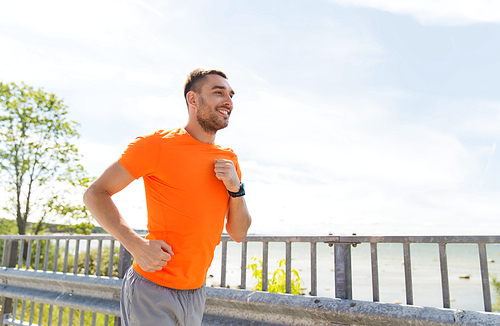 fitness, sport, people, technology and healthy lifestyle concept - smiling young man with heart rate watch running at summer seaside