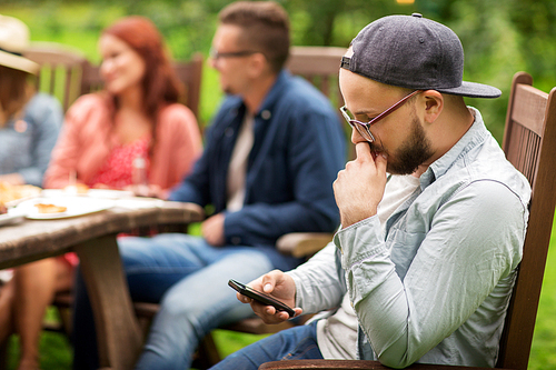leisure, holidays, people and technology concept - young man texting on smartphone and friends having dinner at summer garden party