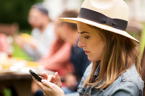 leisure, holidays, people and technology concept - close up of young woman or teenage girl texting on smartphone and friends having dinner at summer garden party