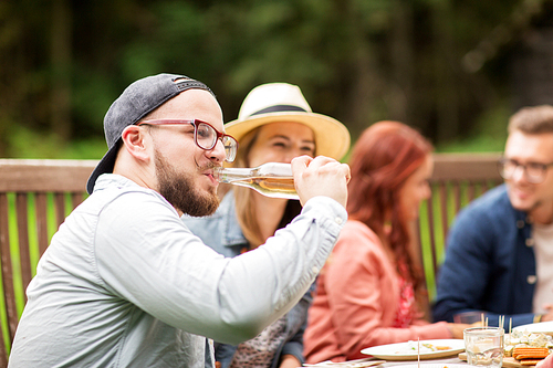 leisure, holidays, eating, people and food concept - happy man drinking beer from bottle at dinner with friends at summer garden party