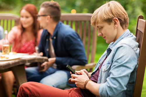 leisure, holidays, people and technology concept - young man texting on smartphone and friends having dinner at summer garden party
