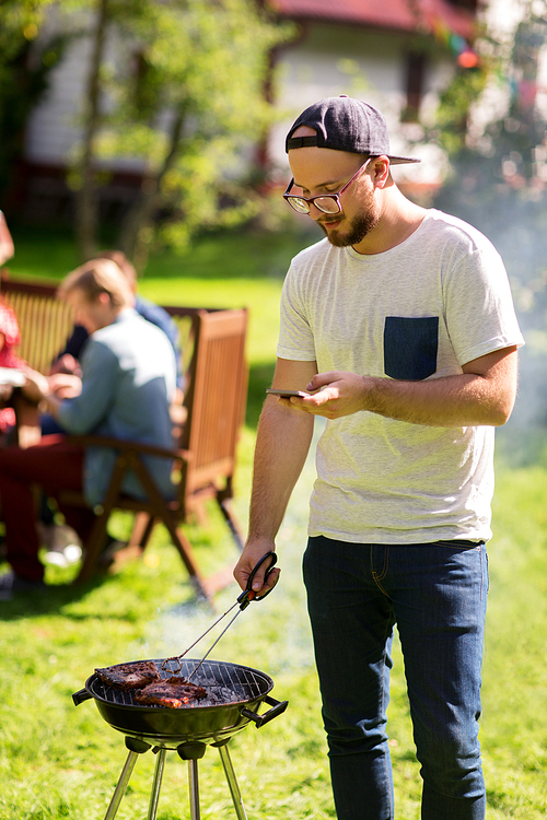 leisure, food, people, technology and holidays concept - man cooking meat on barbecue grill and texting message on smartphone at summer outdoor party