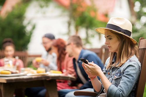 leisure, holidays, people and technology concept - young woman or teenage girl texting on smartphone and friends having dinner at summer garden party