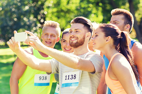 fitness, sport, friendship, technology and healthy lifestyle concept - group of happy sportsmen friends with racing badge numbers taking selfie smartphone outdoors