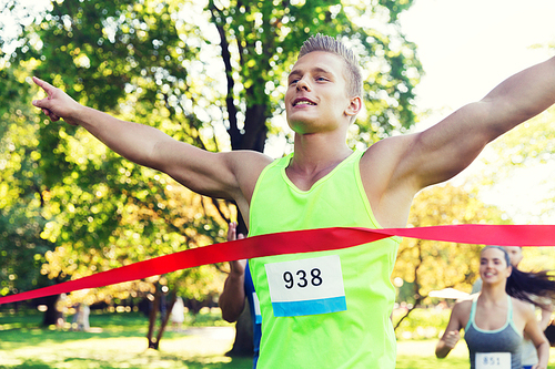 fitness, sport, victory, success and healthy lifestyle concept - happy man winning race and coming first to finish red ribbon over group of sportsmen running marathon with badge numbers outdoors