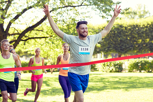 fitness, sport, victory, success and healthy lifestyle concept - happy man winning race and coming first to finish red ribbon over group of sportsmen running marathon with badge numbers outdoors