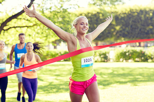 fitness, sport, victory, success and healthy lifestyle concept - happy woman winning race and coming first to finish red ribbon over group of sportsmen running marathon with badge numbers outdoors