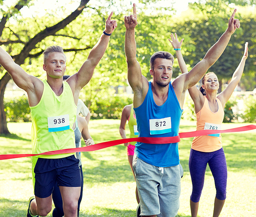 fitness, sport, victory, success and healthy lifestyle concept - happy man winning race and coming first to finish red ribbon over group of sportsmen running marathon with badge numbers outdoors