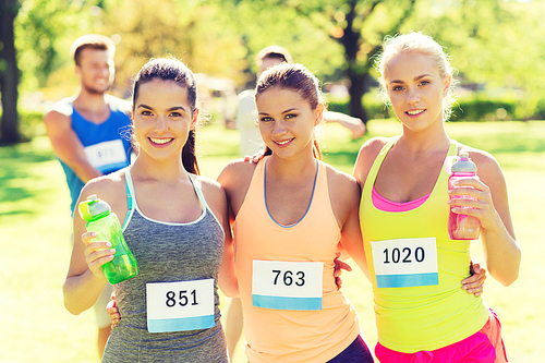 fitness, sport, friendship, marathon and healthy lifestyle concept - happy young sporty women with racing badge numbers and water bottles outdoors