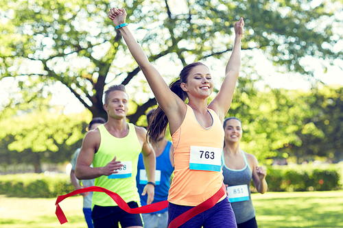 fitness, sport, victory, success and healthy lifestyle concept - happy woman winning race and coming first to finish red ribbon over group of sportsmen running marathon with badge numbers outdoors