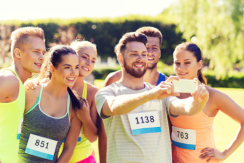 fitness, sport, friendship, technology and healthy lifestyle concept - group of happy sportsmen friends with racing badge numbers taking selfie smartphone outdoors