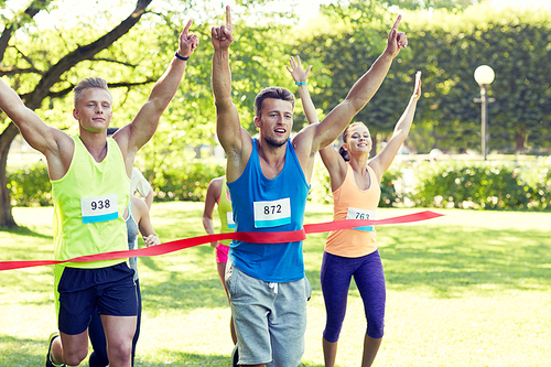 fitness, sport, victory, success and healthy lifestyle concept - happy man winning race and coming first to finish red ribbon over group of sportsmen running marathon with badge numbers outdoors