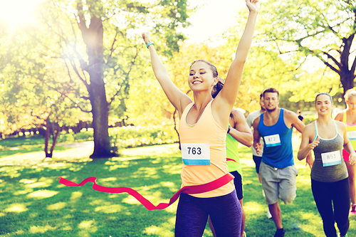 fitness, sport, victory, success and healthy lifestyle concept - happy woman winning race and coming first to finish red ribbon over group of sportsmen running marathon with badge numbers outdoors